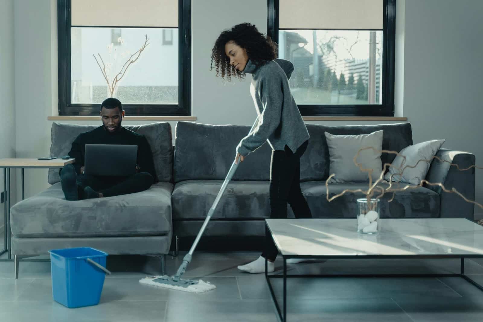A woman mopping floors while the man is busy using laptop.