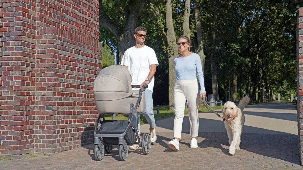 A smiling couple walks on a path, with a man pushing a baby stroller and a woman walking a dog.