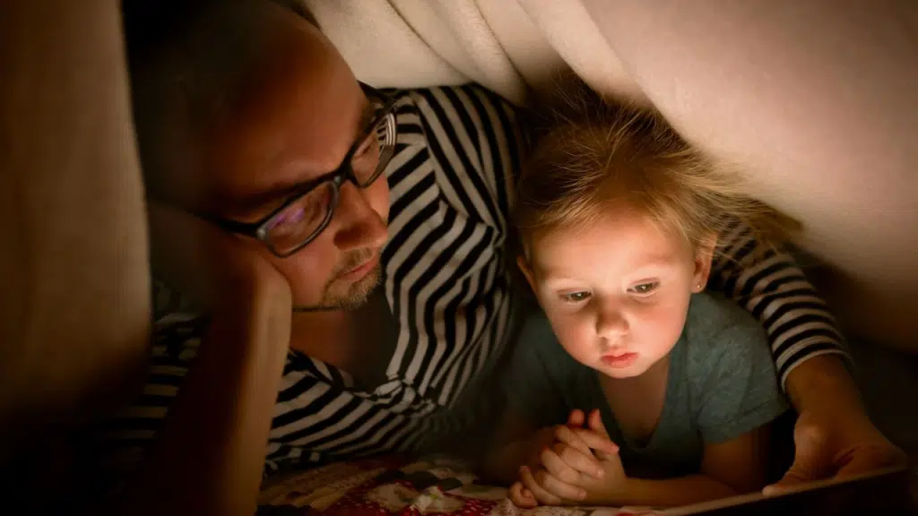 A man and a small child are lying together under a blanket, looking at a tablet with a glowing screen.