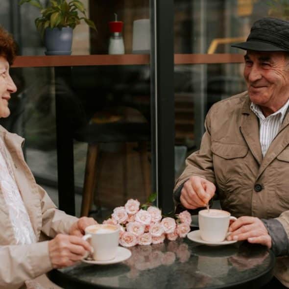 An elderly couple sits at a small table outside a cafe, smiling and enjoying coffee.