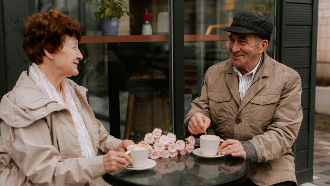 An elderly couple sits at a small table outside a cafe, smiling and enjoying coffee.