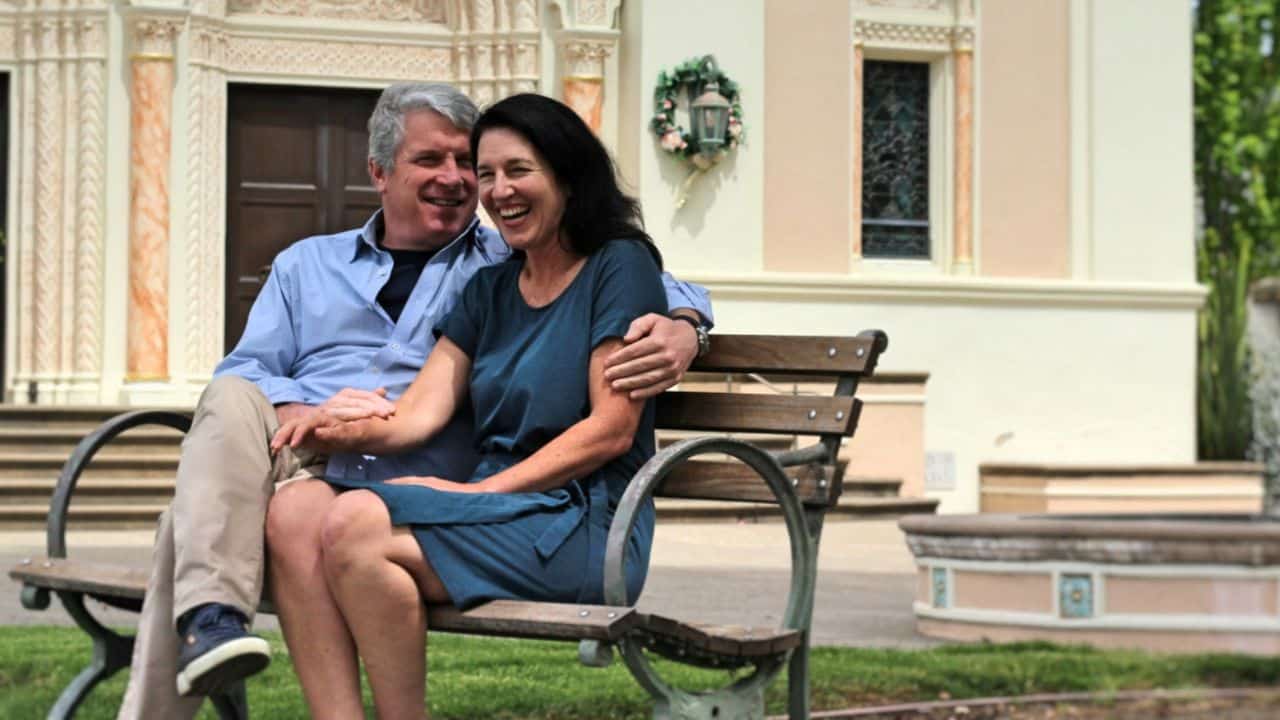 A middle-aged man and woman sit on a park bench and laugh while holding hands.