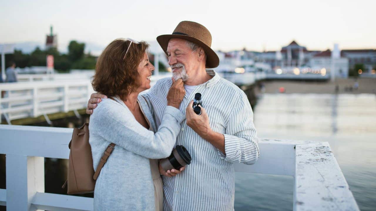 A loving, mature couple smiles at each other on a boardwalk, and the woman touches the man's beard.