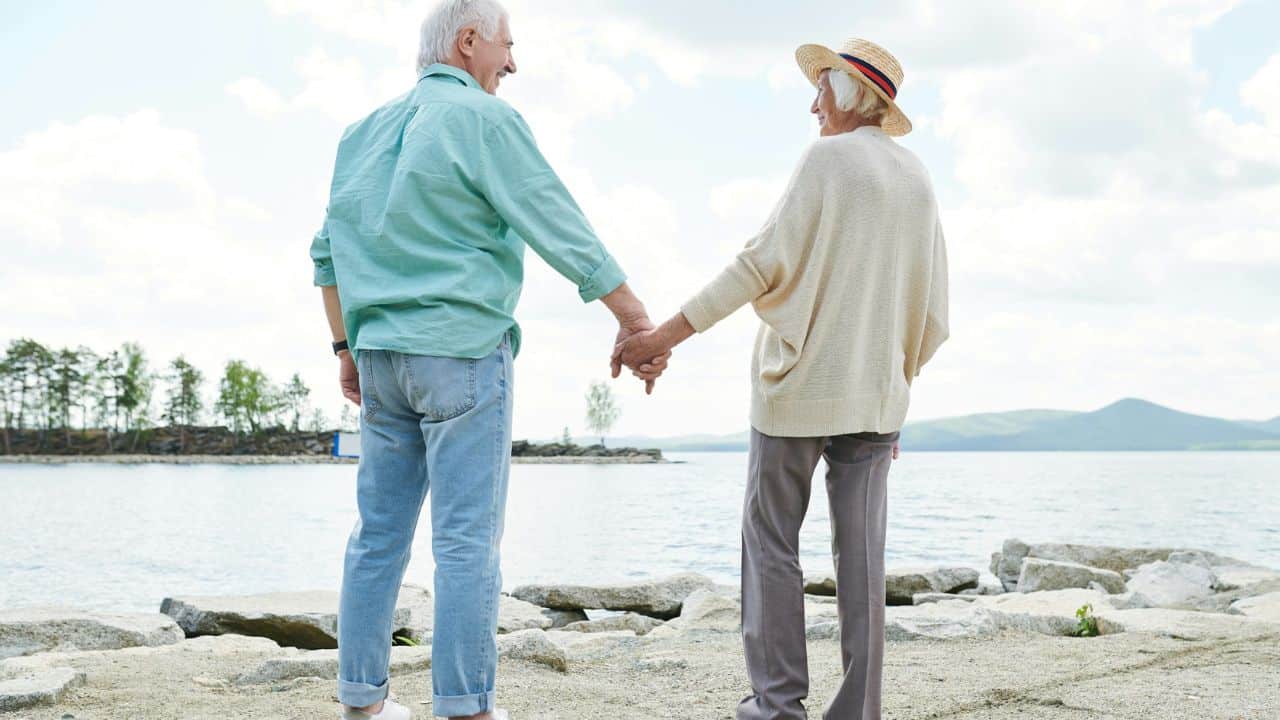 An elderly couple holds hands and looks at each other while standing by a lake.