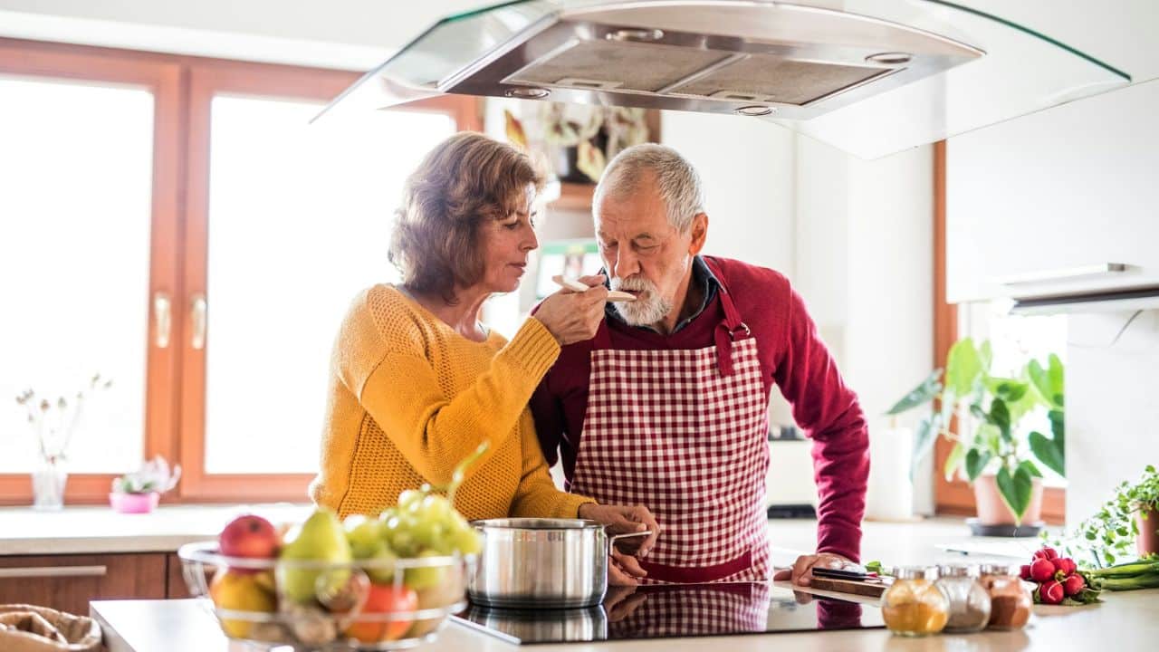 An elderly couple smiles while cooking together in a kitchen. The woman feeds a spoonful of food to the man.