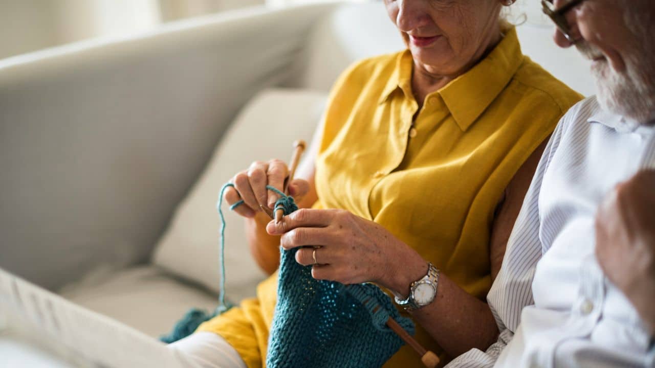 An elderly woman in a yellow shirt knits while sitting next to her husband.