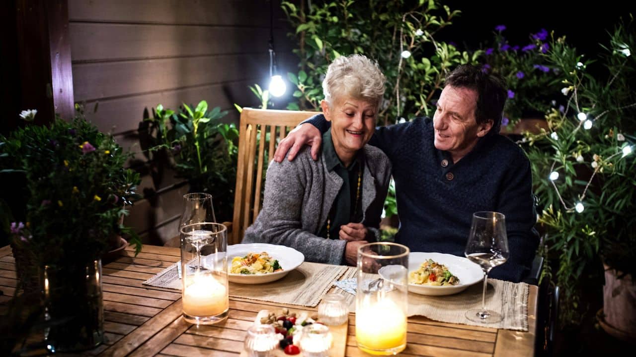 An elderly couple enjoys a romantic dinner on a patio with lights and candles.