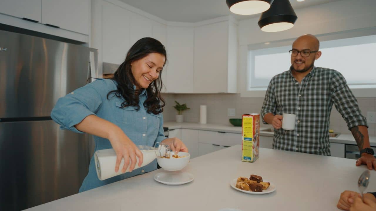 A couple is in a modern white kitchen as the woman pours milk into a bowl of cereal.