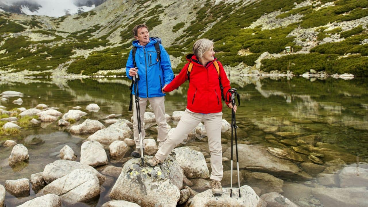 An elderly couple is holding hands while hiking and standing on rocks near a lake.