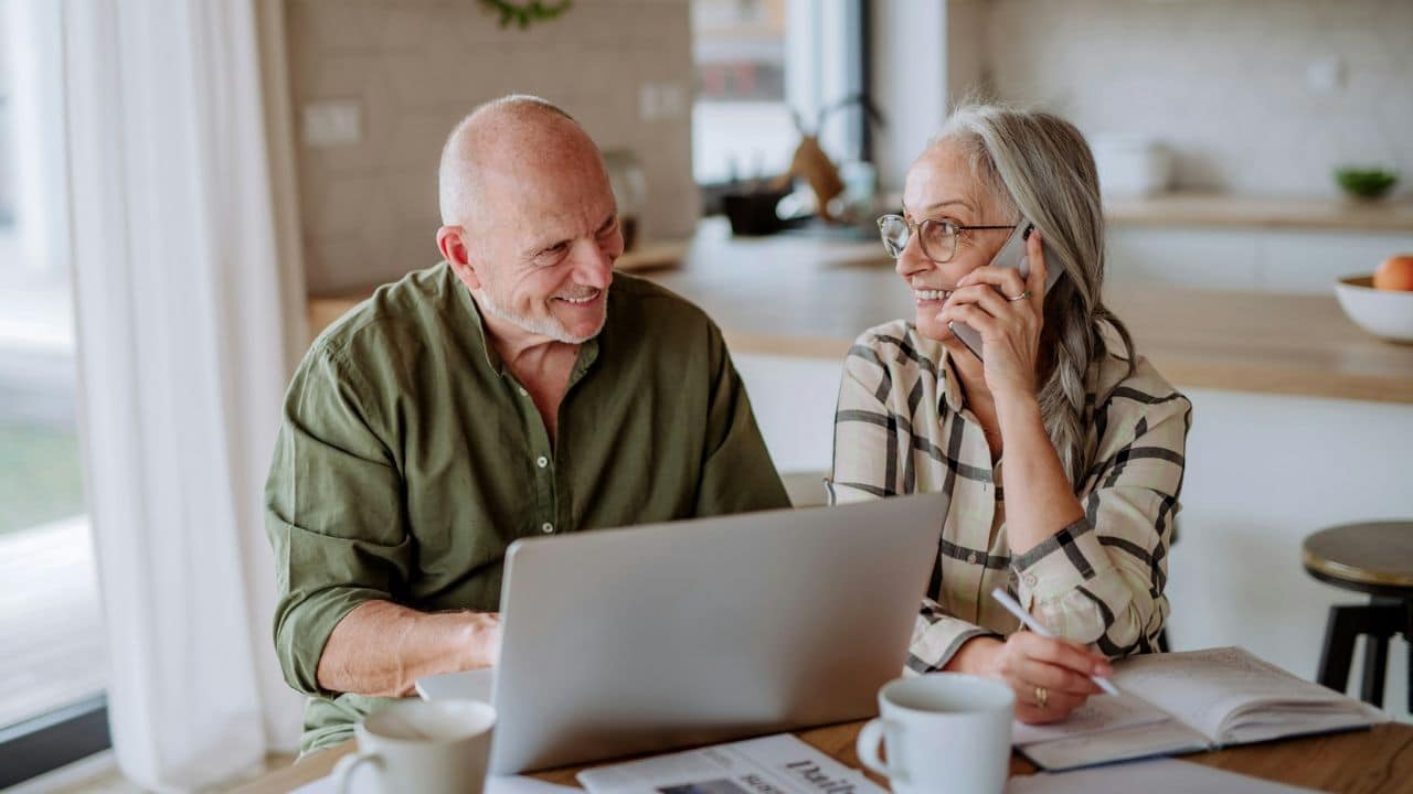 An elderly couple sits at a kitchen table, looking at a laptop and working on finances. The woman is on the phone.