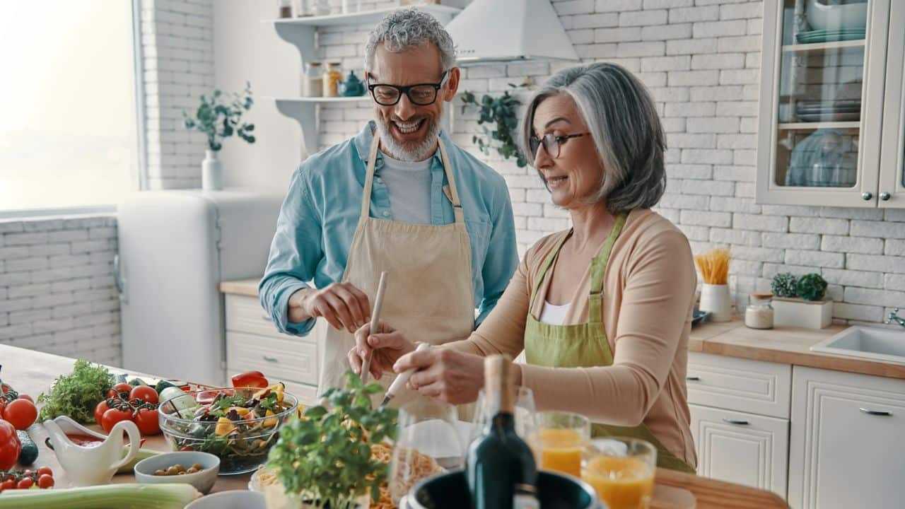 An elderly couple laughs together while cooking in a bright, modern kitchen.