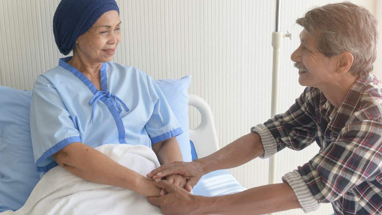 An older man is visiting his sick wife in a hospital bed, holding her hands. She is wearing a headscarf and a light blue patient gown.