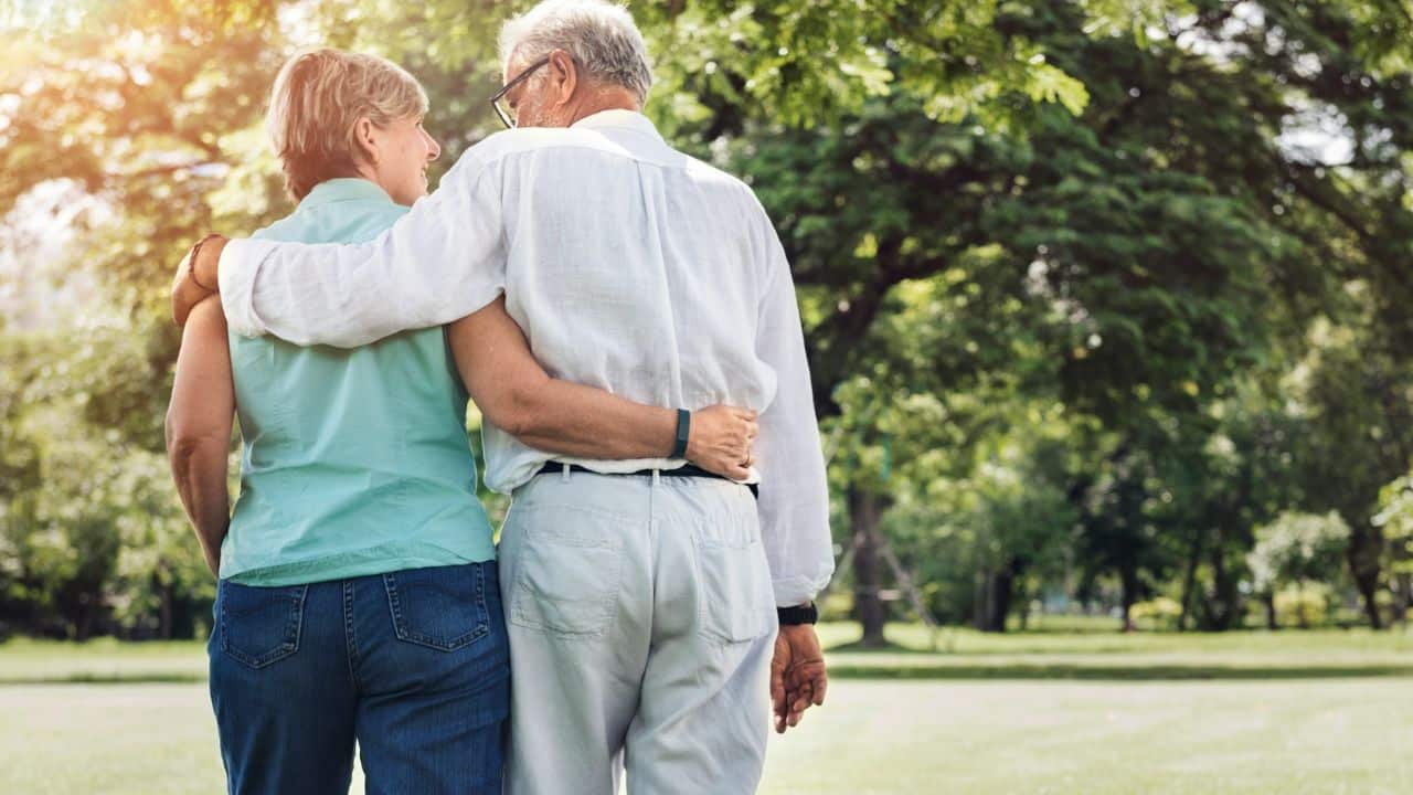 An elderly couple walks with their arms around each other's waist in a park.