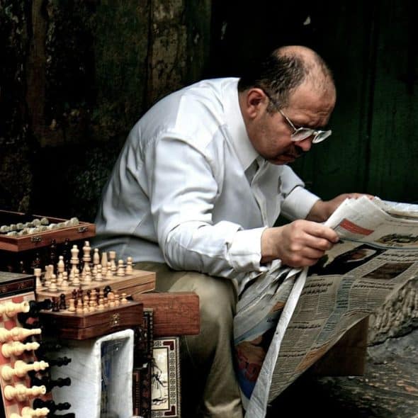 A mature man with glasses reading a large newspaper. A display of chess sets and carved wooden boxes is beside him.