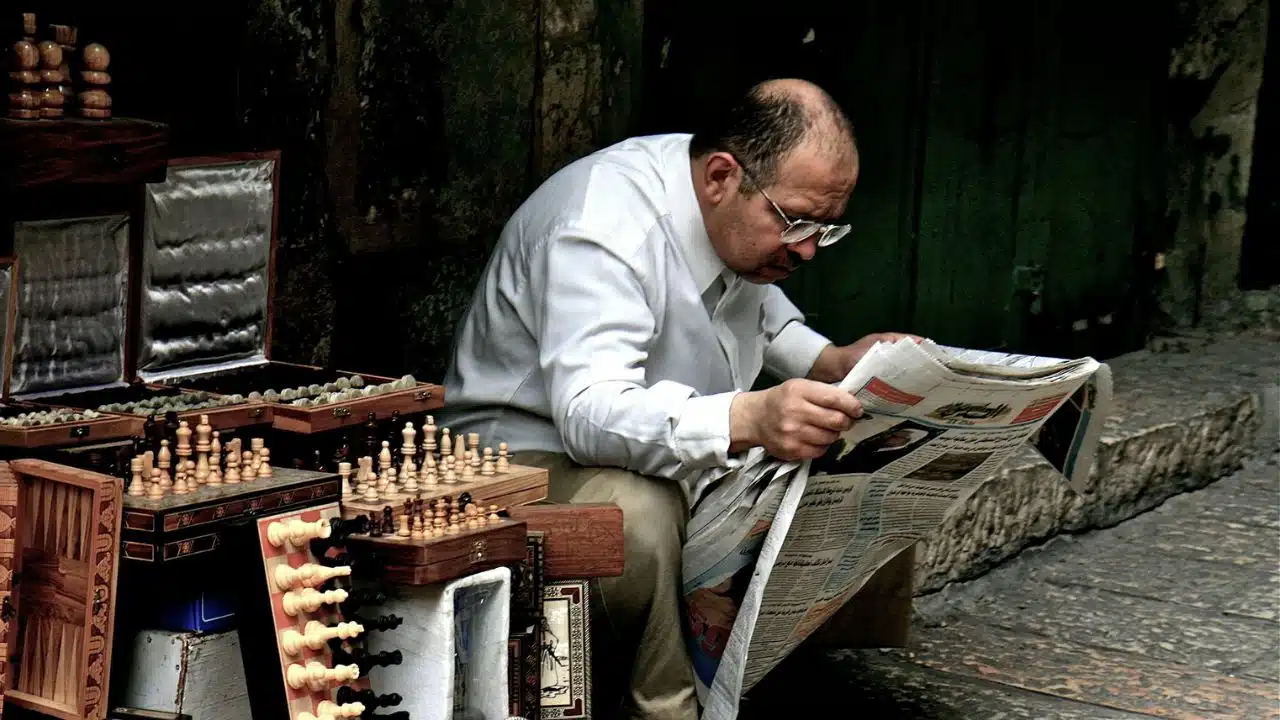 A mature man with glasses reading a large newspaper. A display of chess sets and carved wooden boxes is beside him.