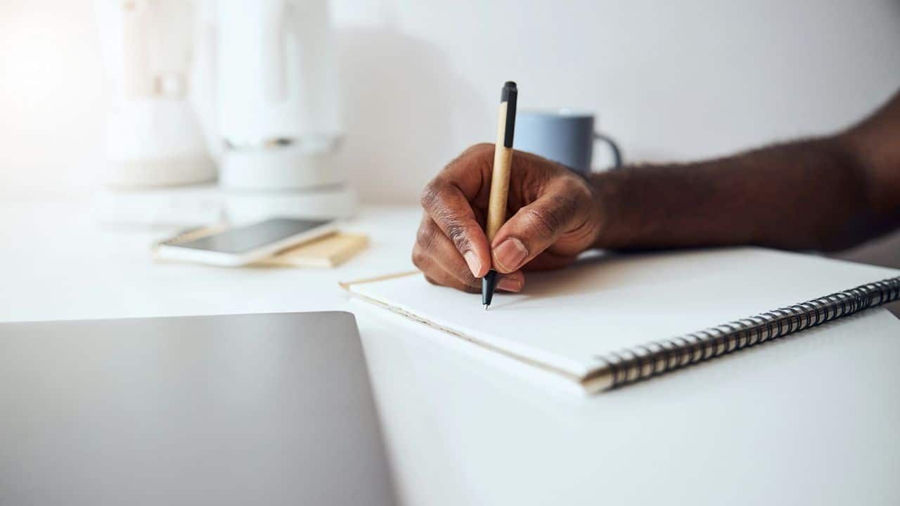 A dark-skinned hand is writing in a spiral notebook with a pen on a white desk.