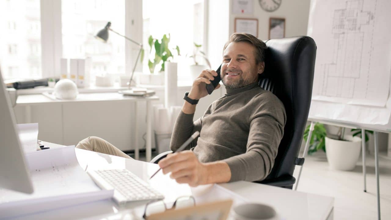 A smiling middle-aged man is sitting in a black leather office chair, talking on his phone while holding a pen.
