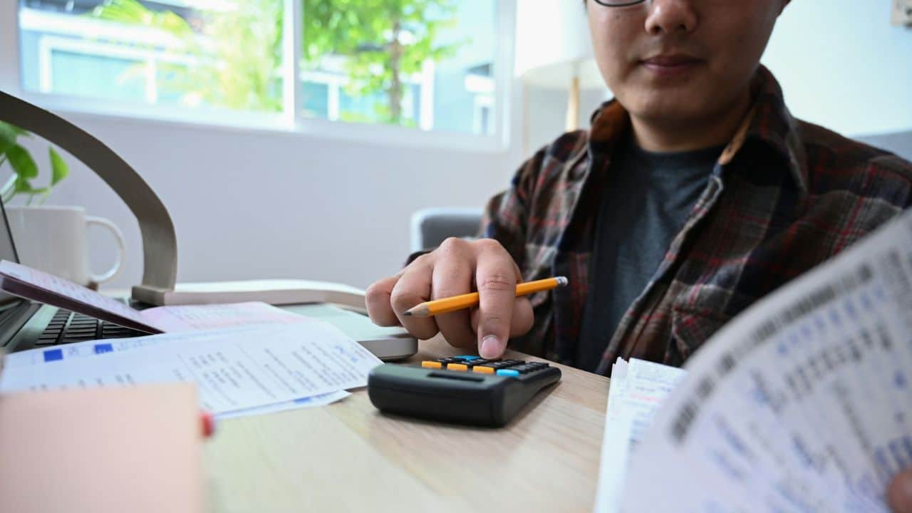 A person wearing glasses and a plaid shirt is using a calculator to pay bills at a wooden table covered in papers.