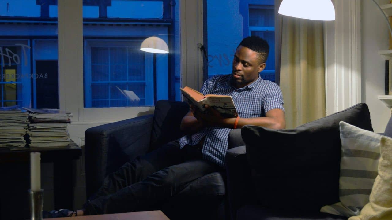 A handsome Black man sits on a dark sofa, intently reading a physical book under a lamp.