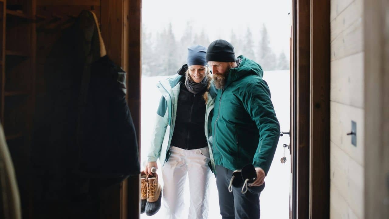 A happy couple in winter gear is taking off their boots and entering a warm-looking cabin.