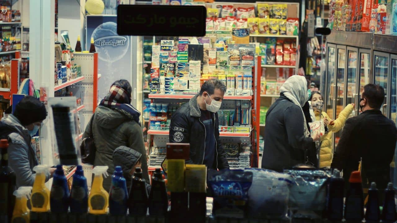 A view through a store window shows several people wearing face masks shopping for groceries and household items.