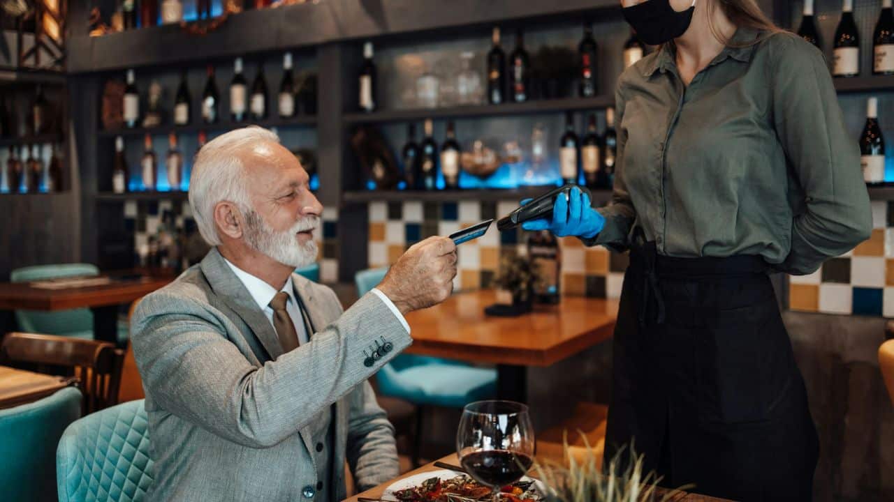 A smiling gentleman is paying for his meal with a credit card at a restaurant. A waitress wearing a face mask and blue gloves holds a payment terminal.