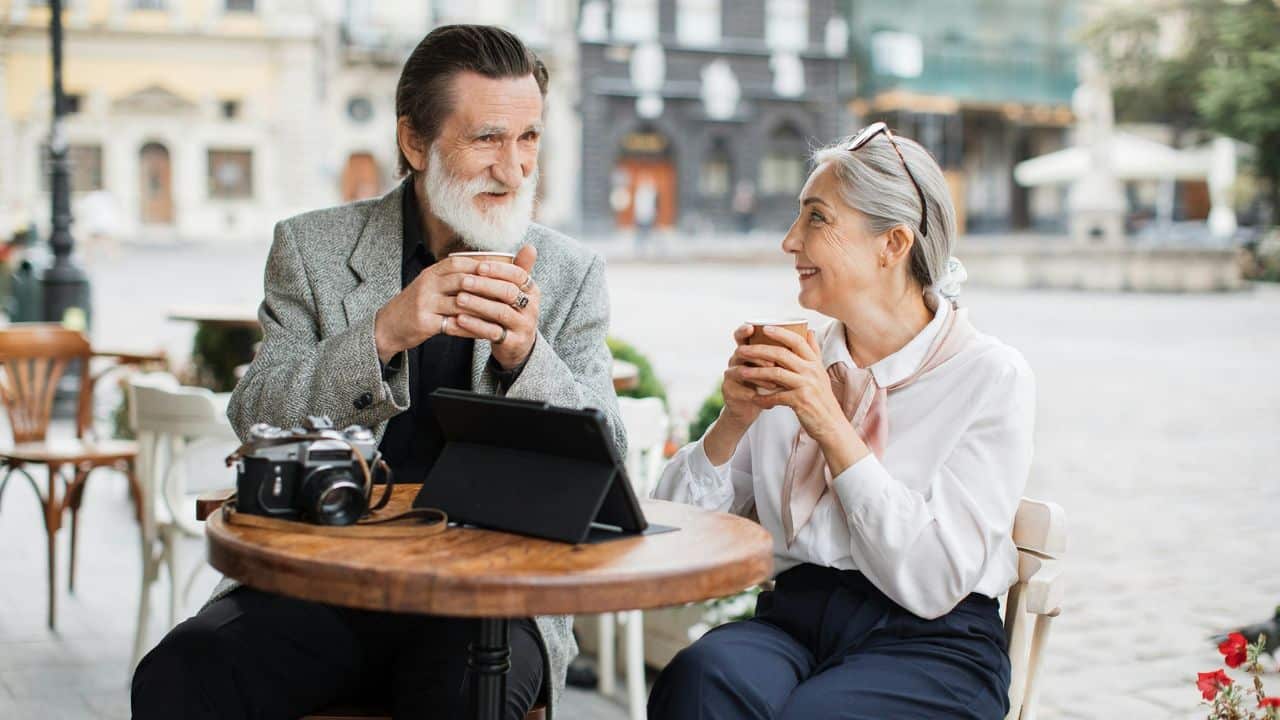 An elderly couple sits at a cafe table on a city street, holding mugs and smiling at each other.