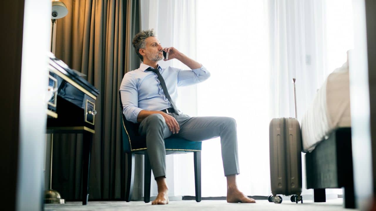 A handsome, mature businessman sits barefoot in a hotel room, talking on a smartphone.