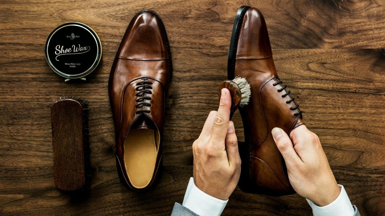 A pair of hands is using a brush to polish a brown leather dress shoe on a wooden table. A tin of shoe wax and another brush are nearby.