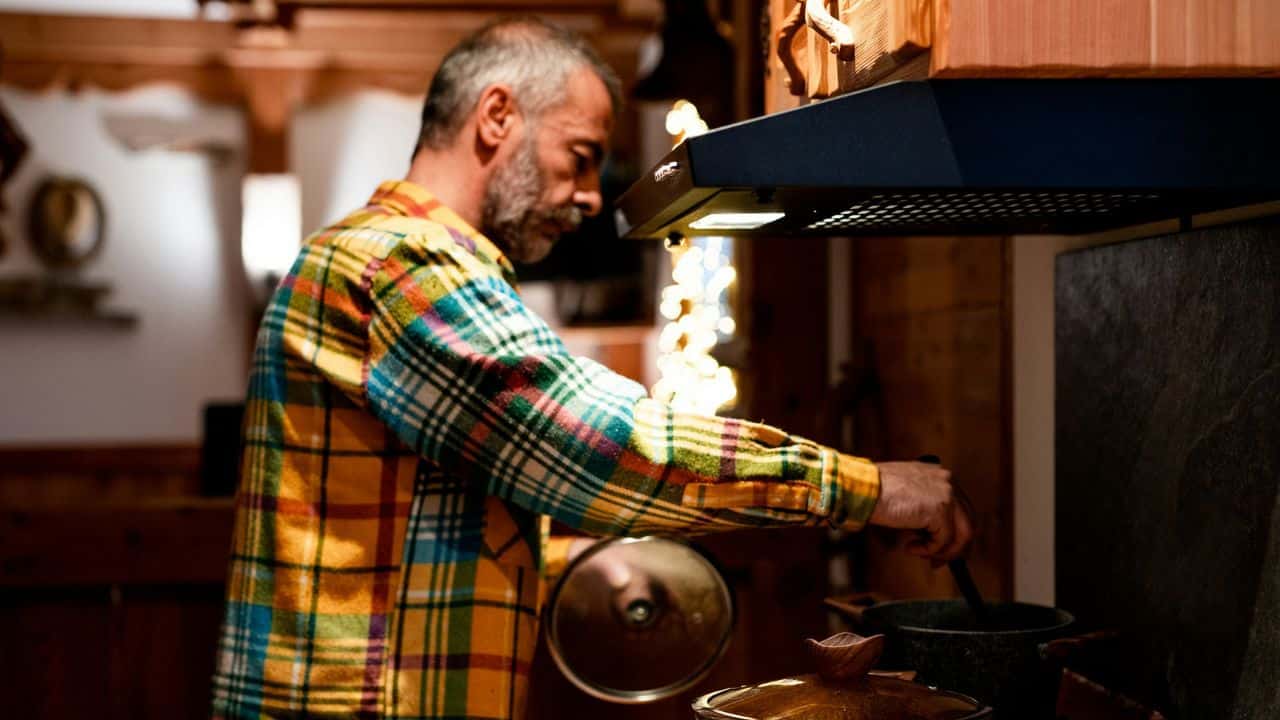 A mature man with a beard is cooking on a stove in a rustic kitchen. He is wearing a colorful plaid shirt, stirring a pot, and holding a lid.
