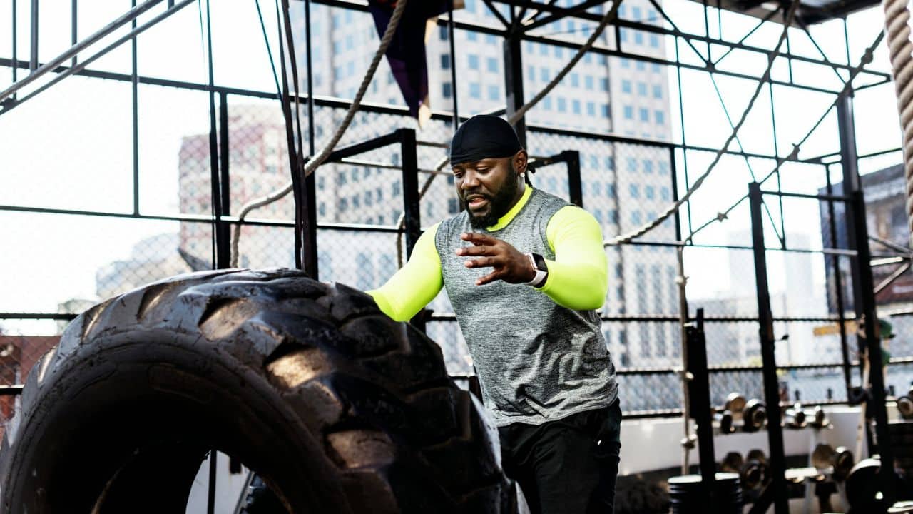 A strong, middle-aged man is pushing a large tire at a gym. He is wearing a black beanie, a grey sleeveless hoodie over a neon green long-sleeve shirt, and is focused on his task.