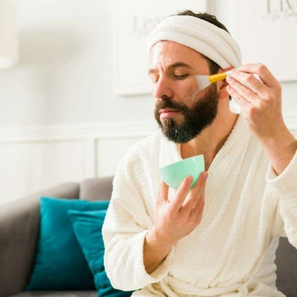 A man with a beard and a white robe is applying a facial mask with a brush.