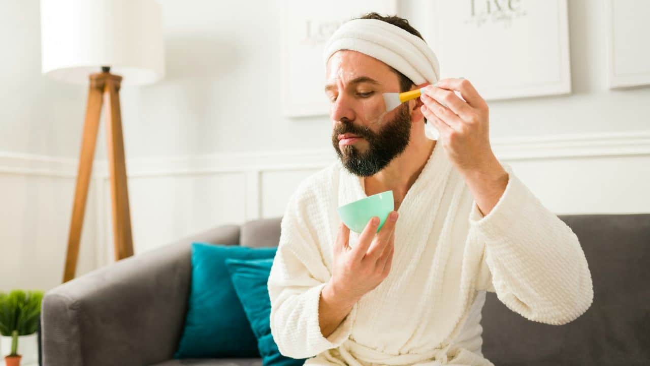 A man with a beard and a white robe is applying a facial mask with a brush.