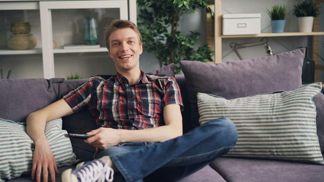 A happy man sits on a couch, holding a remote control and smiling at the camera.