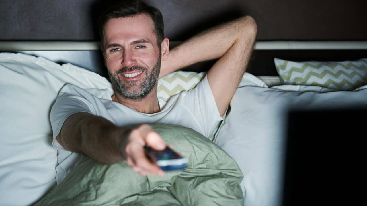 A happy man in bed smiles while holding a remote control and watching television.