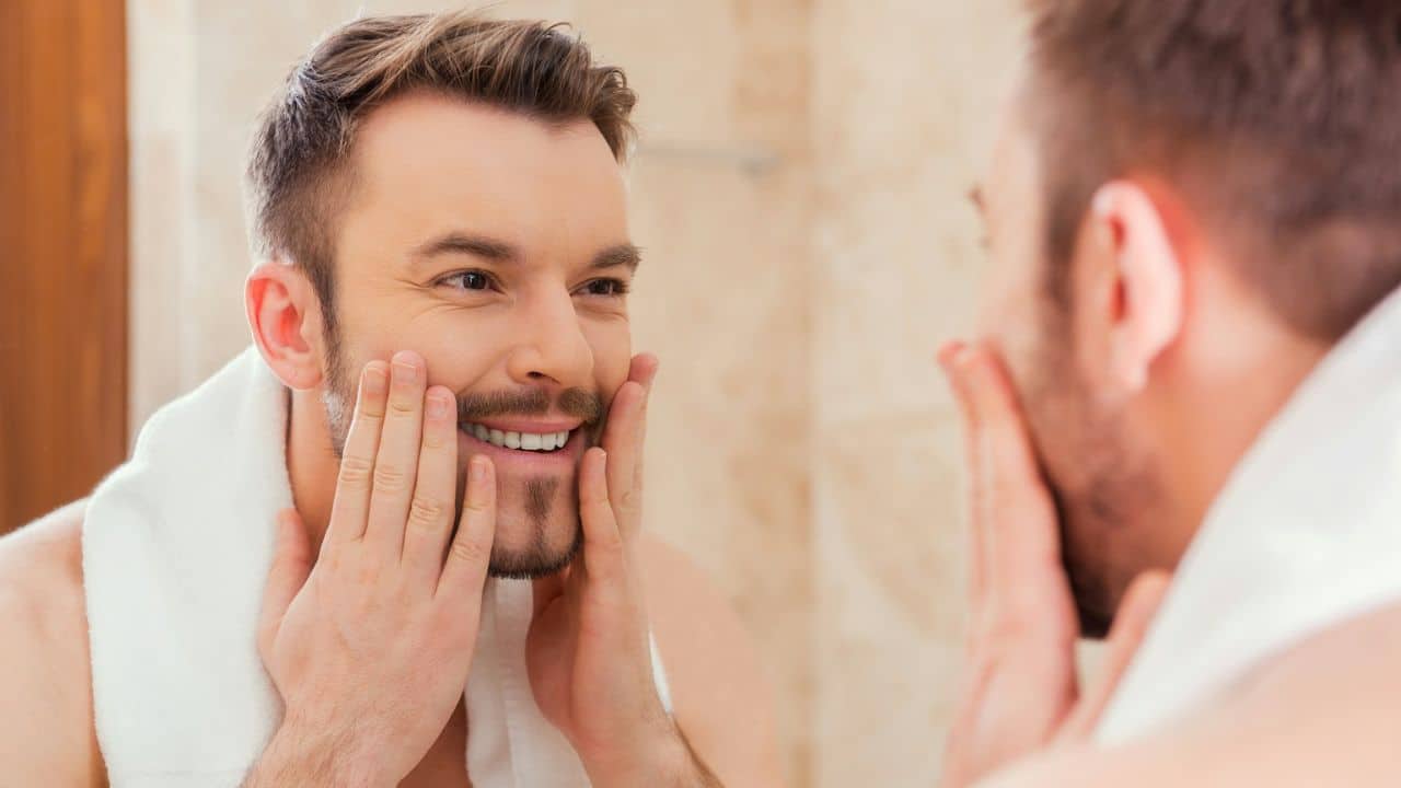 A happy man with a towel around his neck is looking at his reflection while touching his face.