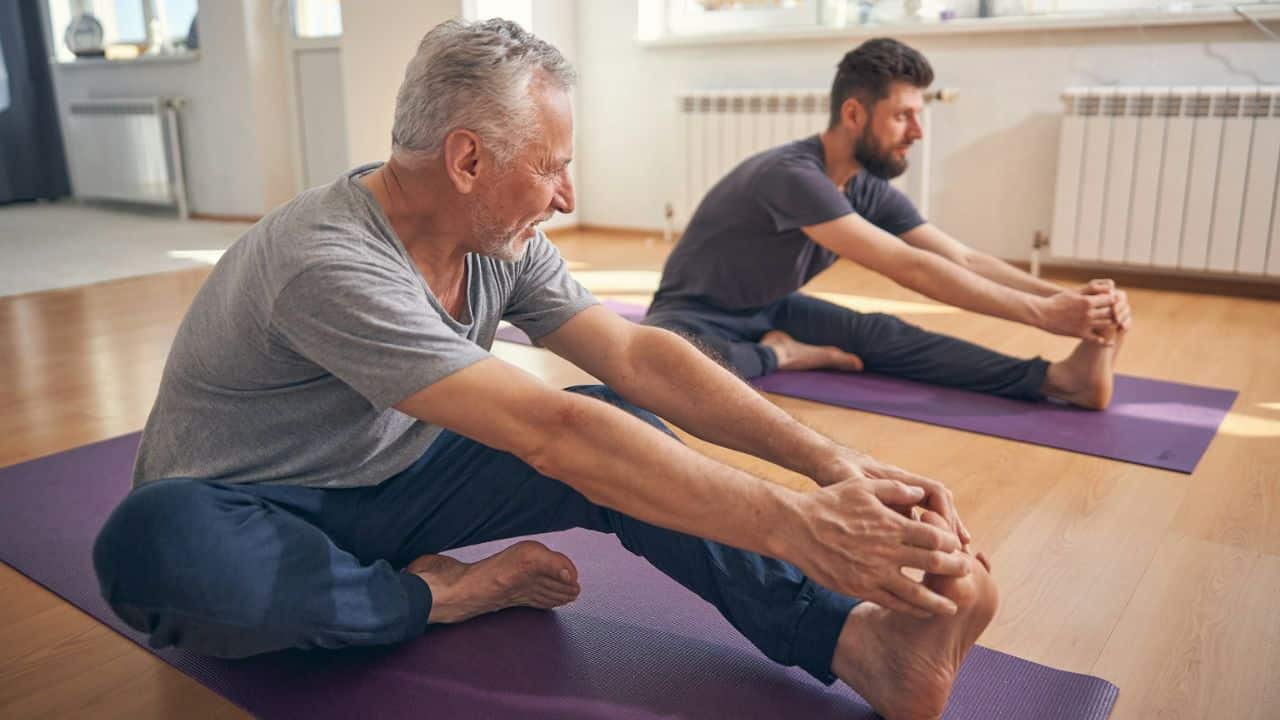 Two men on yoga mats are doing a forward fold and touching their toes.