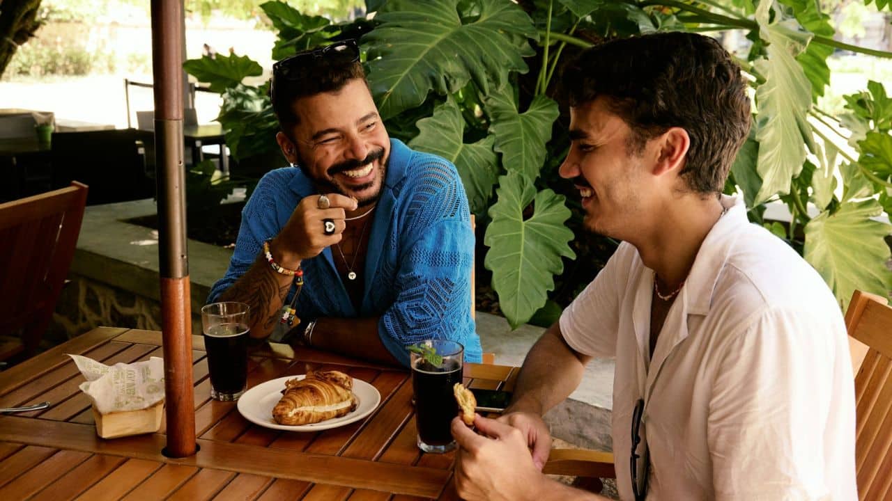 Two men are laughing and talking while sitting at a table with drinks and food.