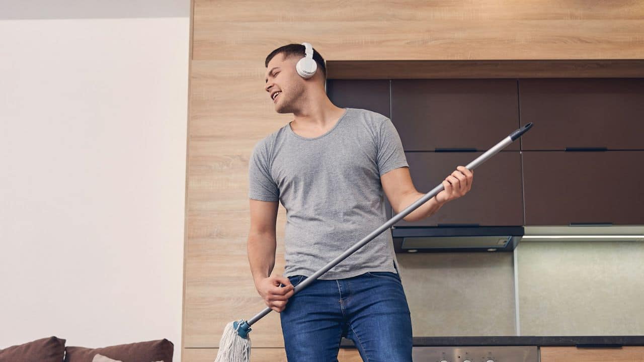A man with headphones on is singing into a mop handle, pretending it's a microphone.