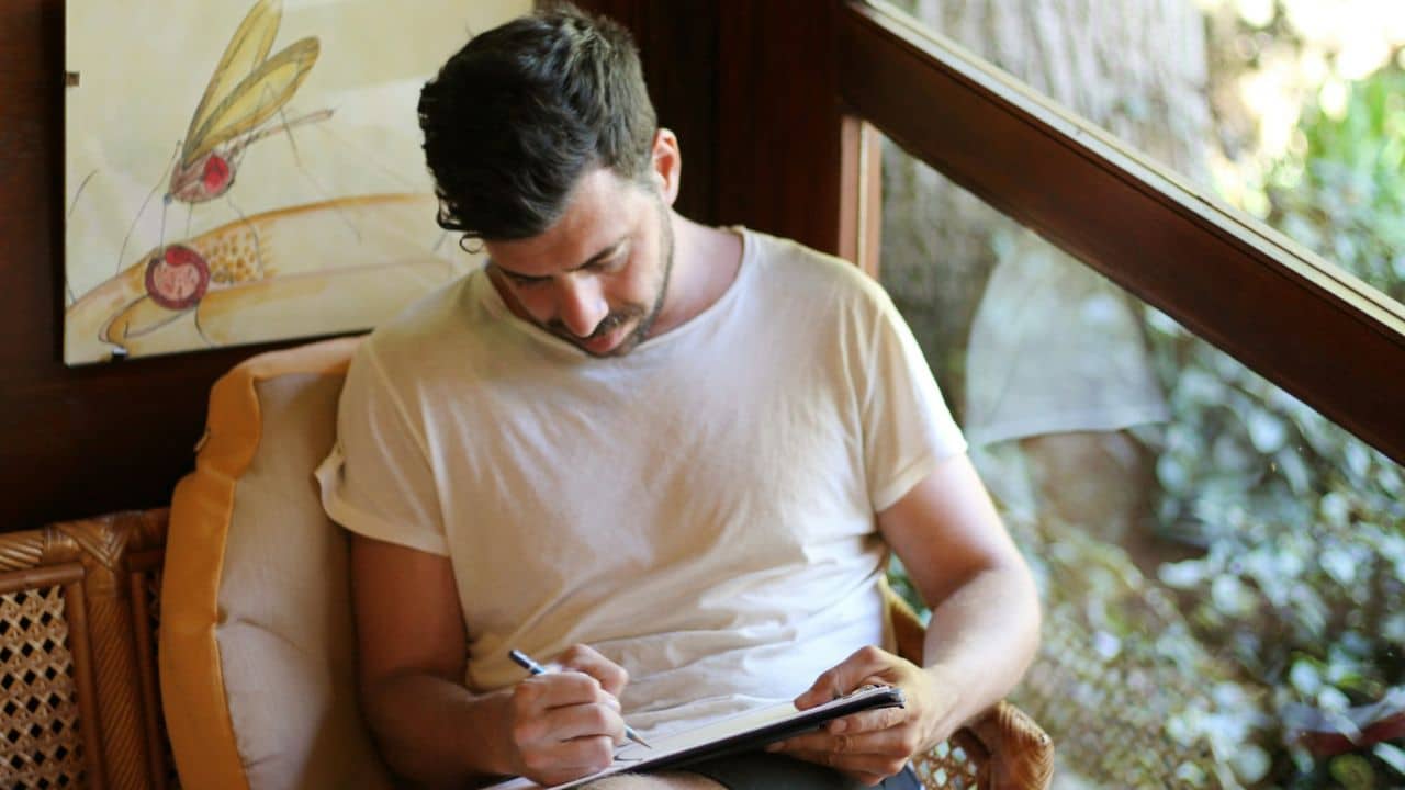 A man in a white T-shirt sits in a wicker chair, writing in a journal.
