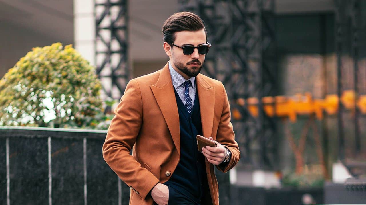 A fashionable man in sunglasses, a tie, and a camel coat is holding a wallet outdoors.