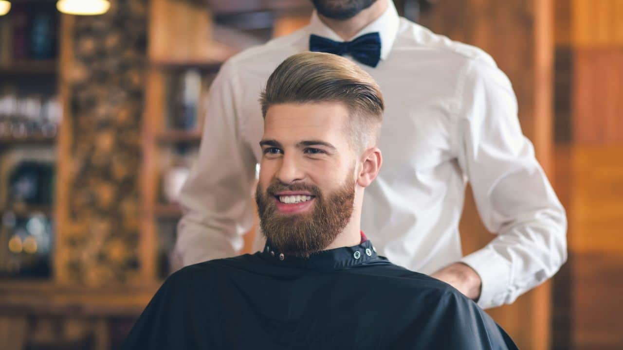 A bearded man in a salon chair is smiling as a barber stands behind him.