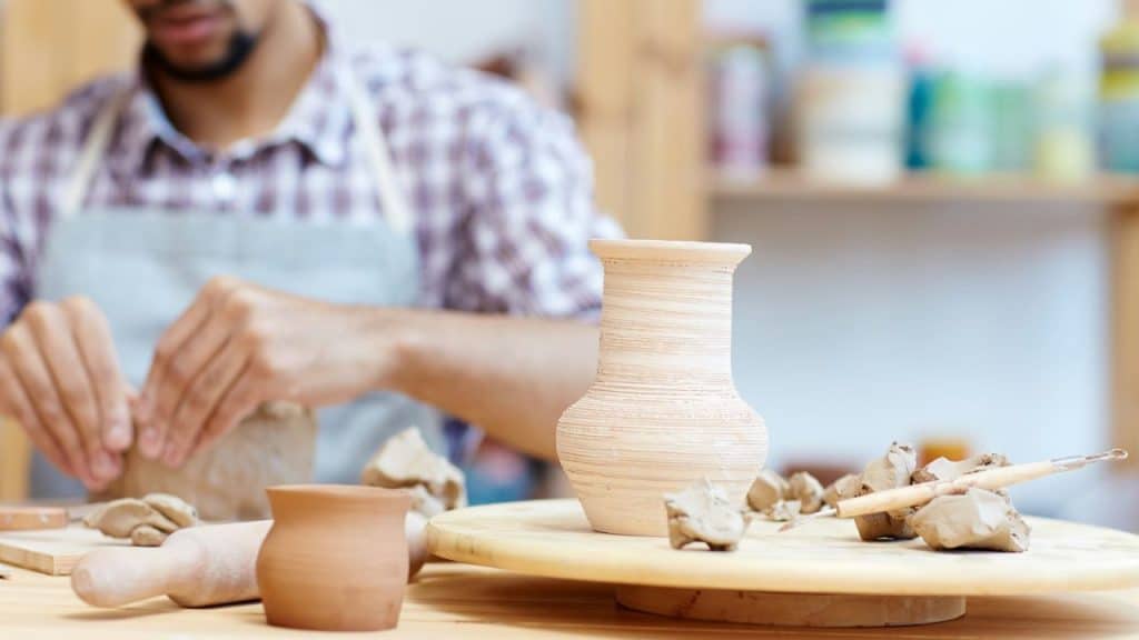 A man is making pottery in a studio with a finished vase on a table.