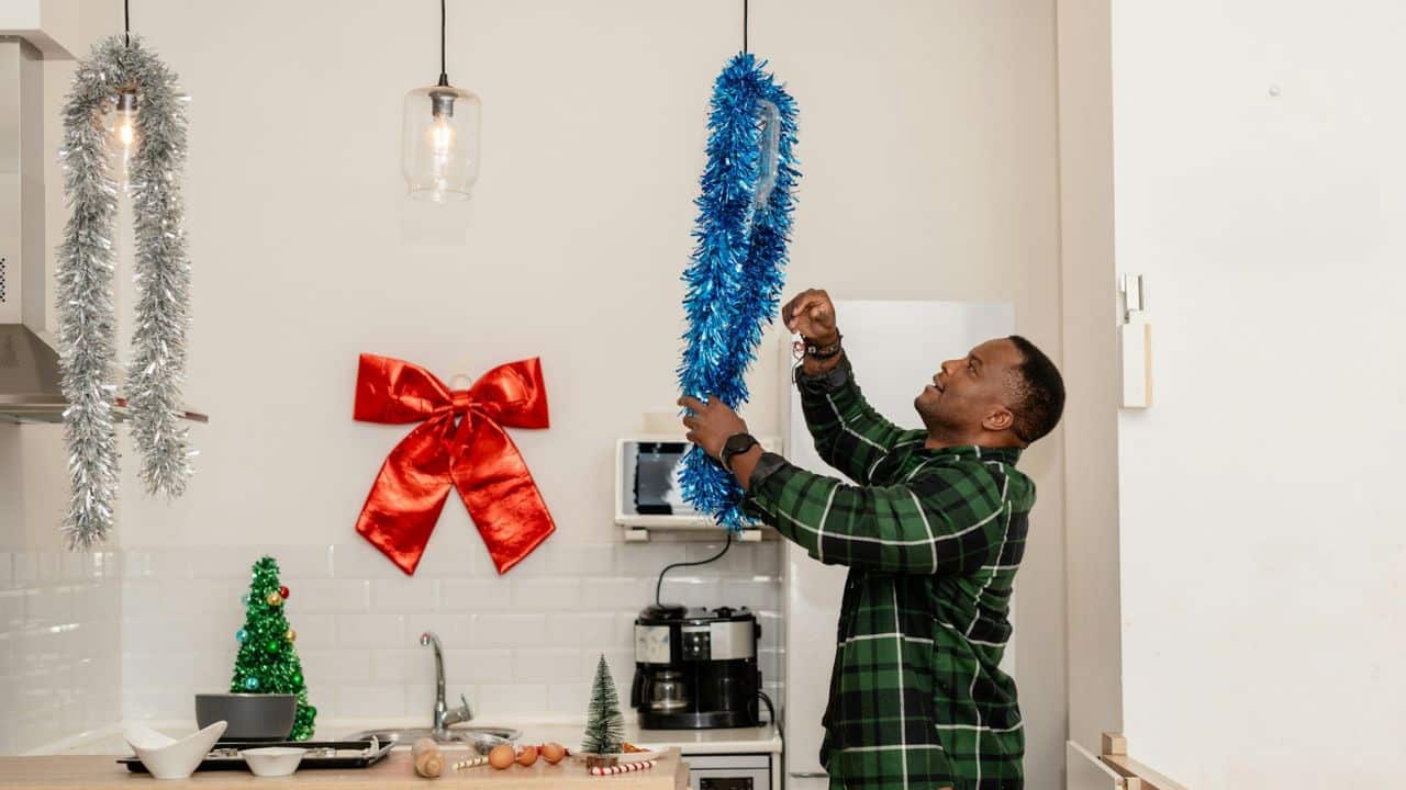 A man in a green flannel shirt is hanging blue tinsel in a kitchen decorated for the holidays.