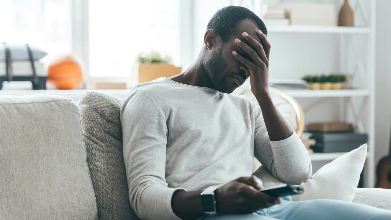 A man on a couch holds his face with one hand and a remote control with the other.