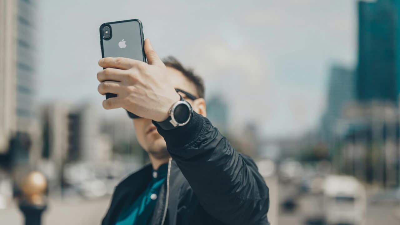 A man is taking a selfie with his iPhone, wearing sunglasses and a jacket.