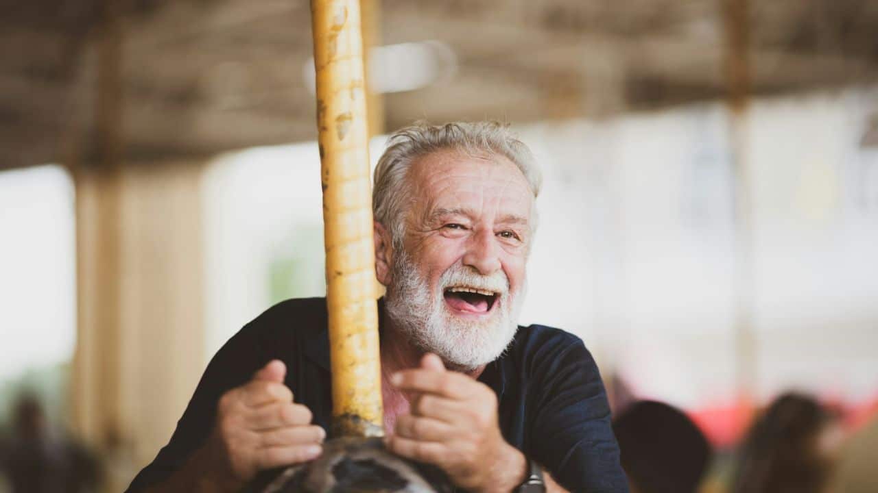 A senior man with a white beard is joyfully laughing, riding on a carousel.