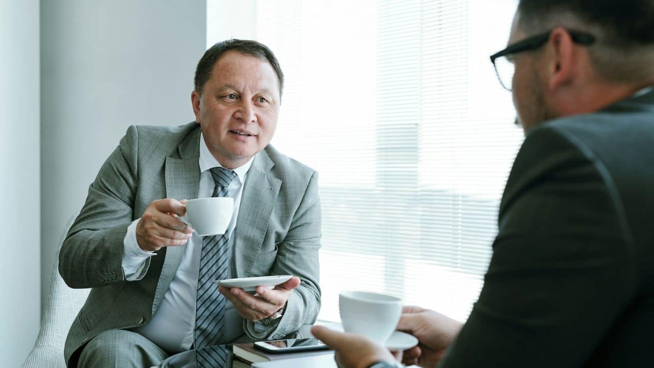 Two men in suits are sitting together, drinking from coffee cups, and talking.
