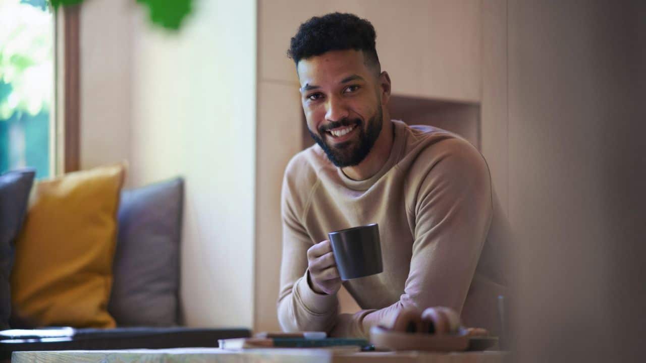 A handsome, happy man with a beard is smiling at the camera and holding a coffee mug.