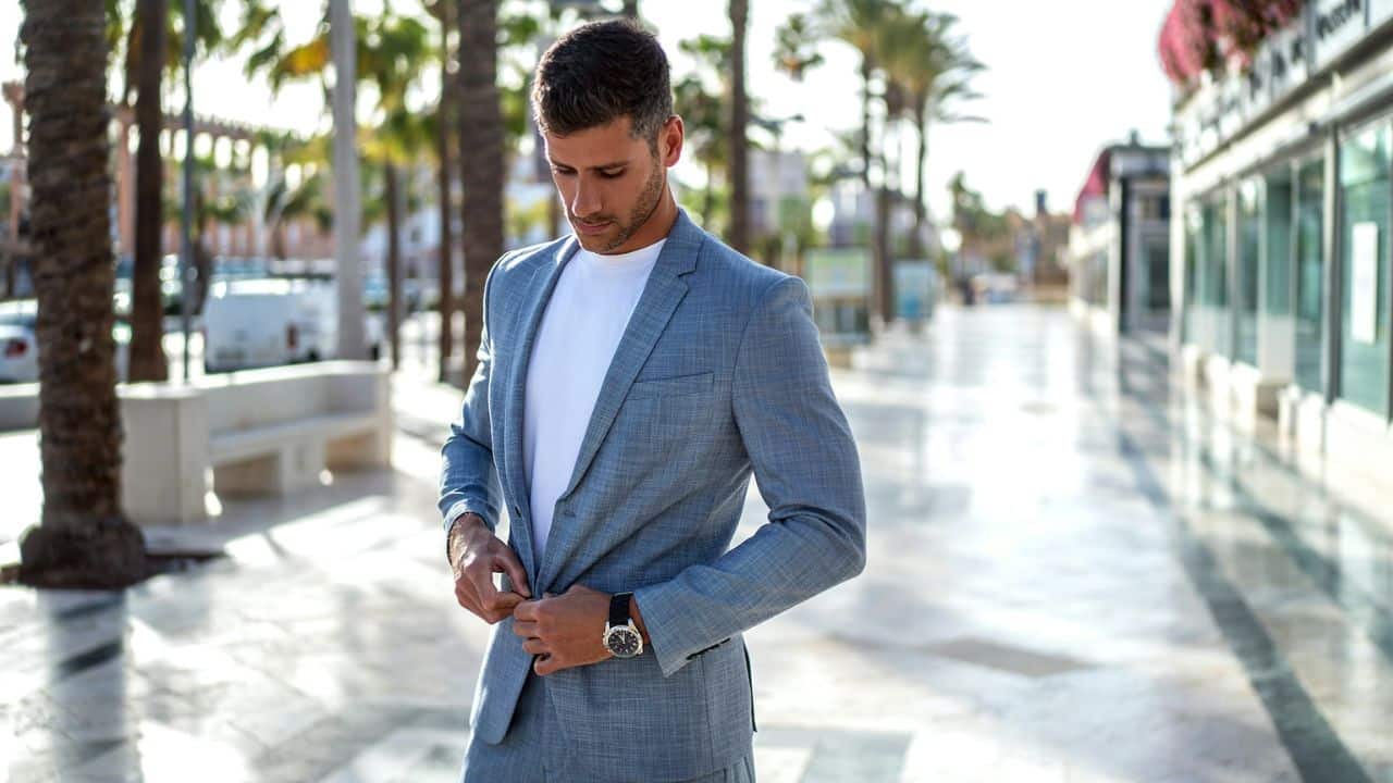 A man in a light blue suit and white t-shirt buttons his jacket while standing on a walkway.